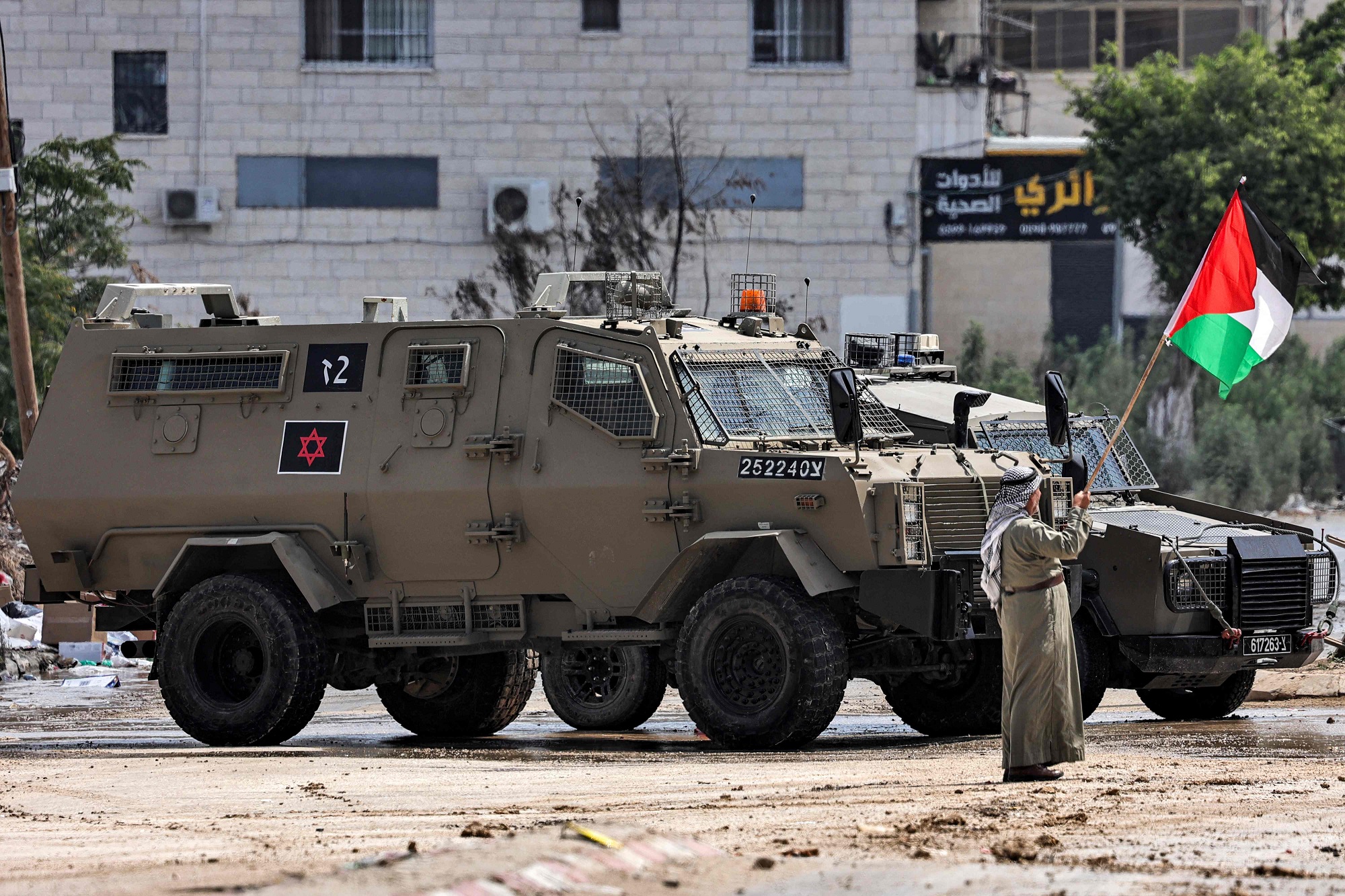 An elderly man stands with a Palestinian flag before Israeli occupation army armored vehicles deployed at the entrance of the Tulkarm camp for Palestinian refugees, during an ongoing Israeli raid in Tulkarm in the occupied West Bank on August 22, 2024. (Photo by Zain JAAFAR / AFP)